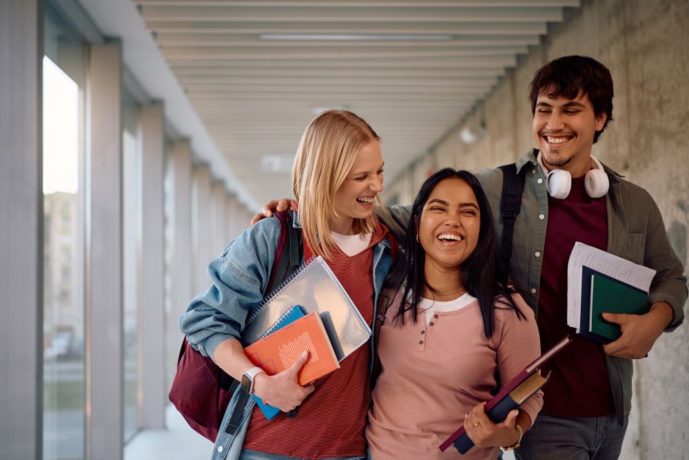 Three students walk through a campus together, carrying textbooks and laughing